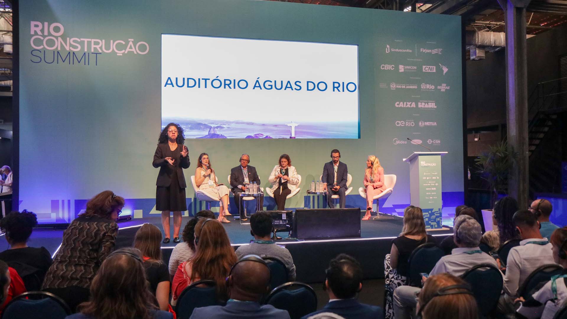 Palco em primeiro plano com cinco debatedores no palco, três mulheres e dois homens